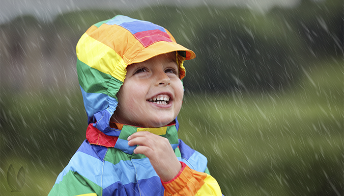 A child is wearing a rainbow colored rain jacket while standing in the rain.