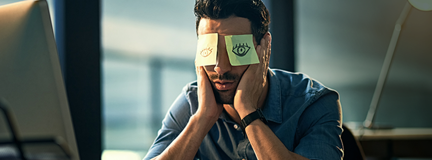 A man at work is pretending to be awake by drawing fake eyes on sticky notes